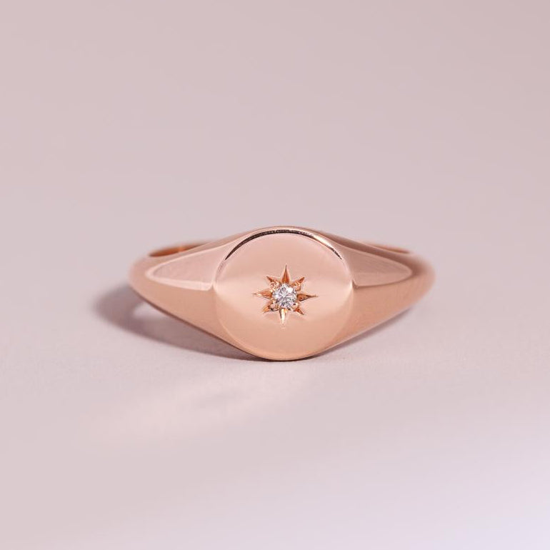A solid 9ct rose gold midi monogram ring with a round polished face, flat to the finger, set with central natural round brilliant cut diamond displayed against a light background.